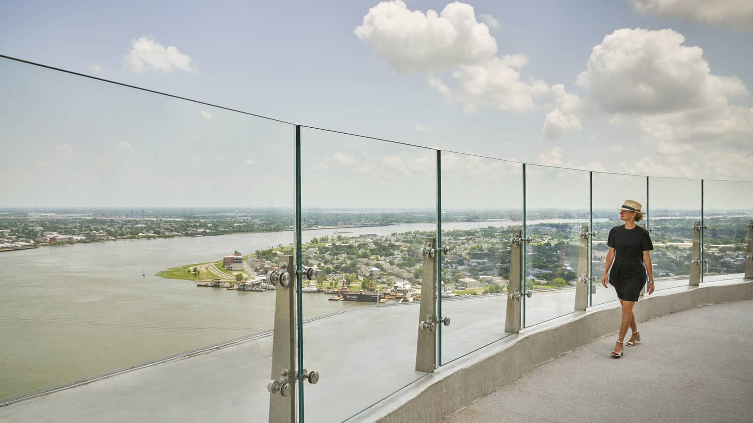 A woman in black clothes walking along a glass wall of an observation deck looking out at a river.