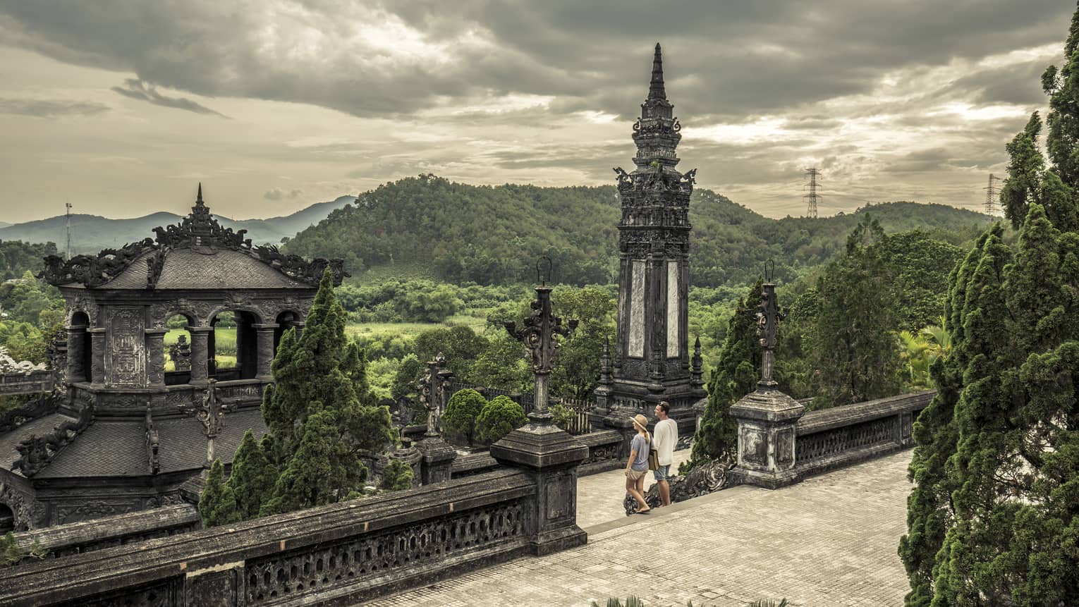 Man and woman walk down steps of historic Imperial City of Hue with stone towers, rolling hills