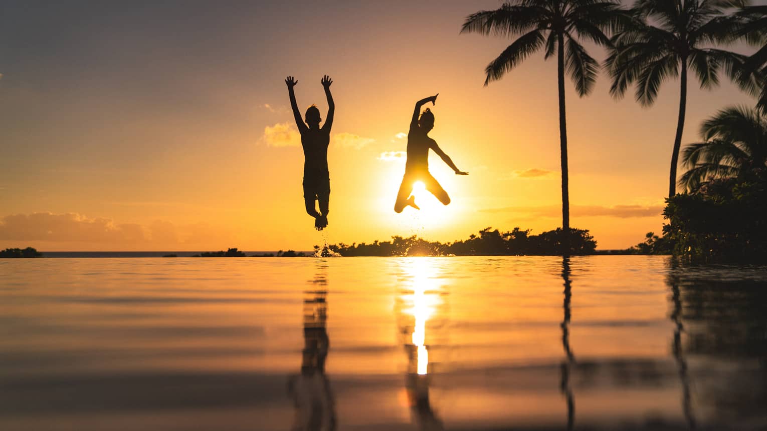 Silhouette of two children jumping above the water, sunset and palm trees in background 