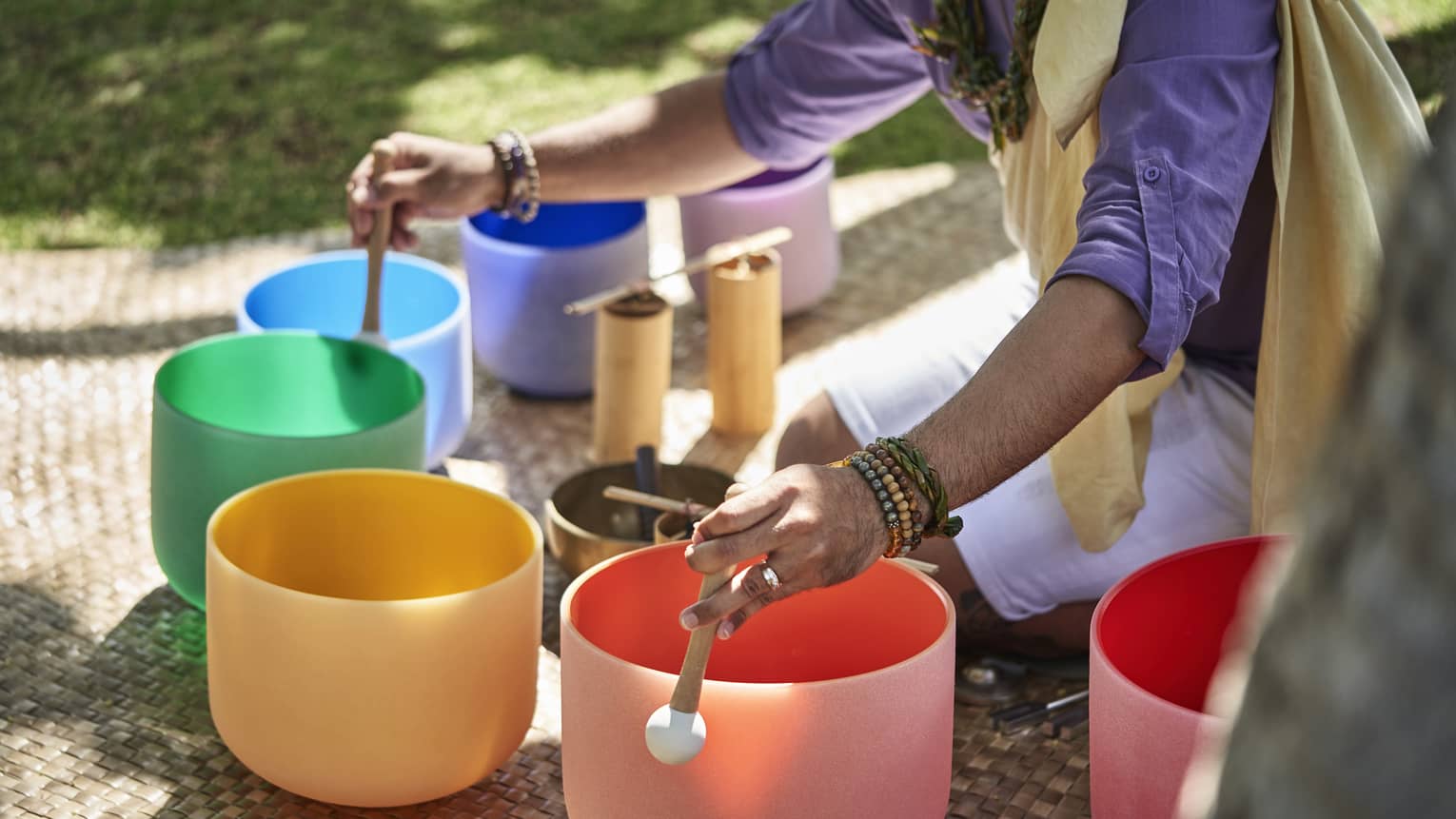 Close up of a man's hands using mallets to play a row of colourful crystal sound bowls