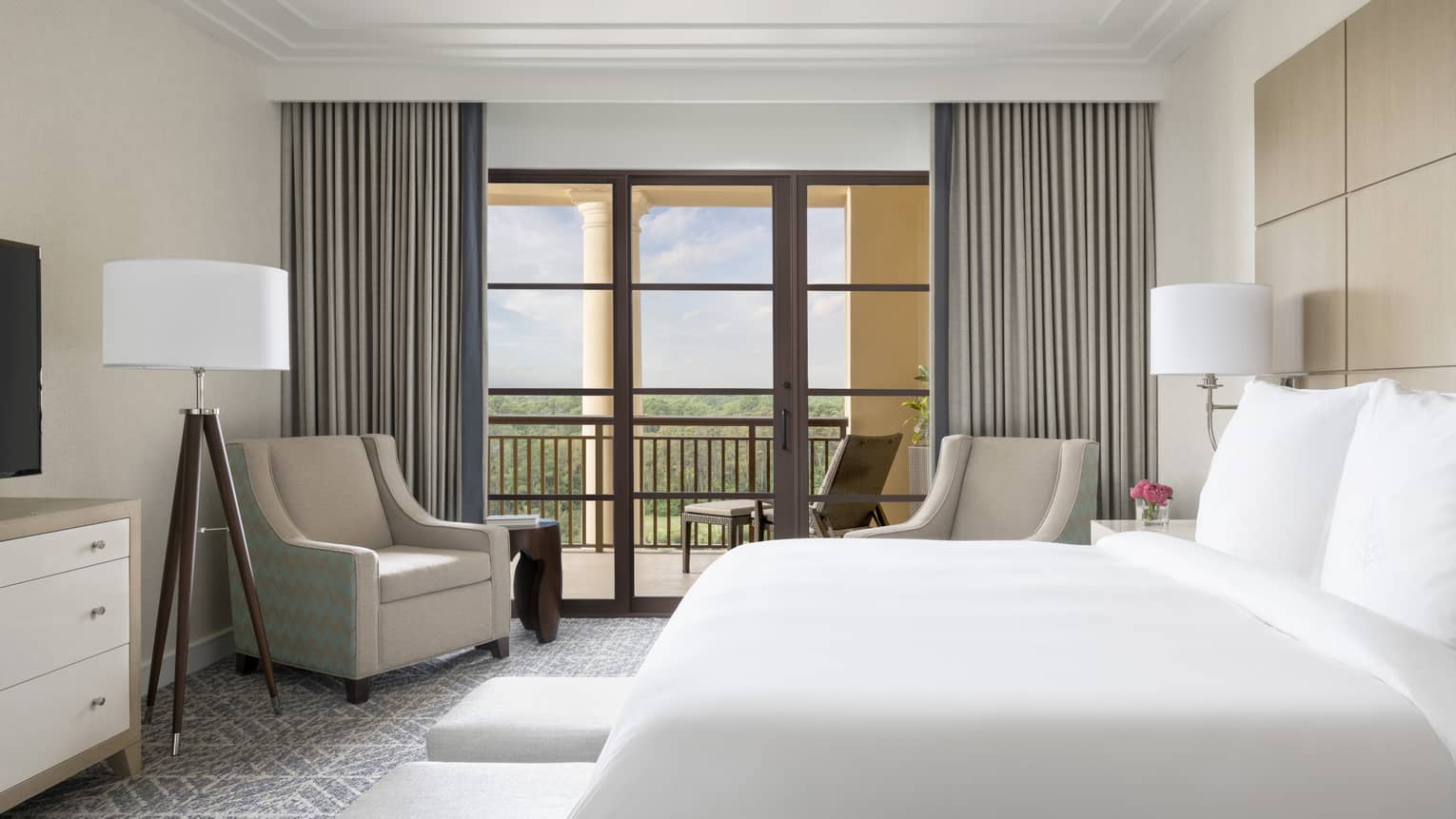 Hotel room with white king bed, two beige arm chairs, grey curtains, walk-out balcony