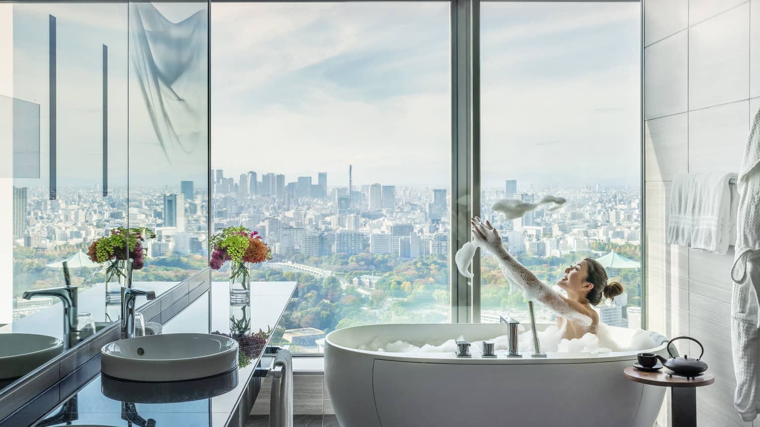 Woman in soaking tub in guest bathroom, floor-to-ceiling windows with city views