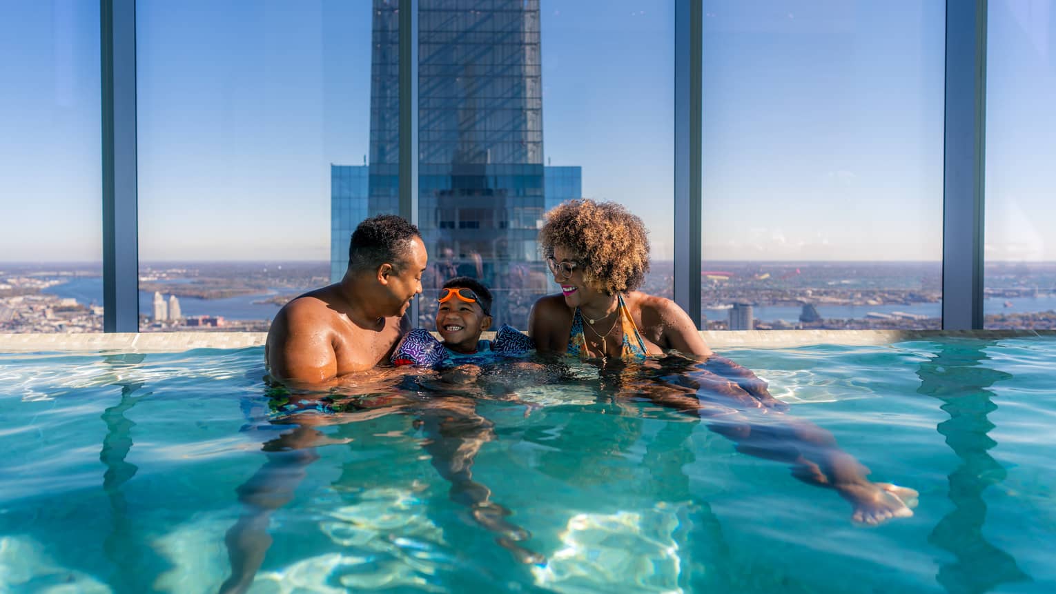 A father, mother and son in a pool surrounded by large windows looking out at a city skyline.