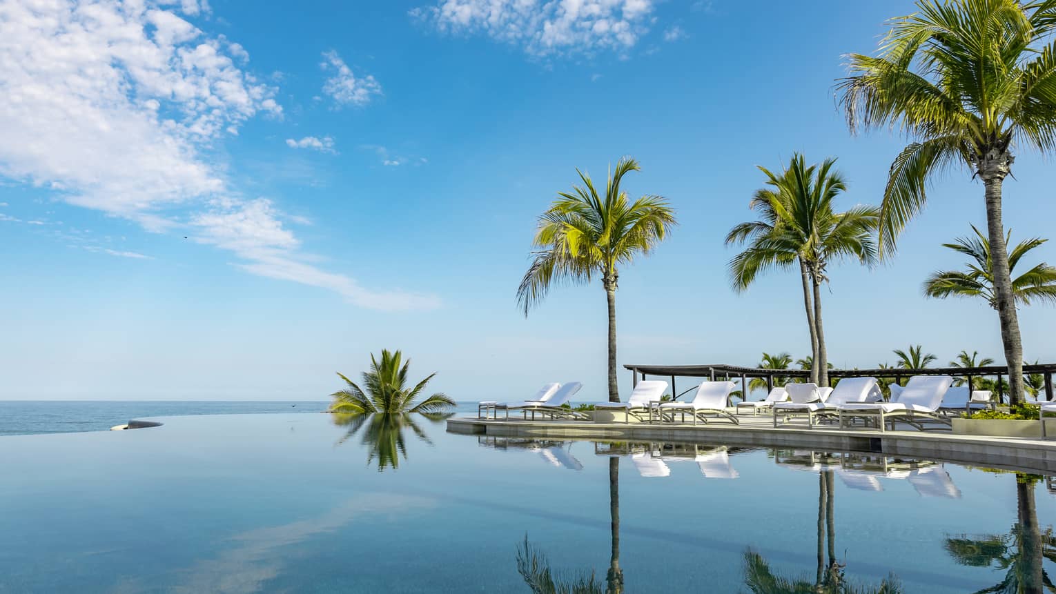 A calm infinity pool reflects the blue sky, clouds and palm trees