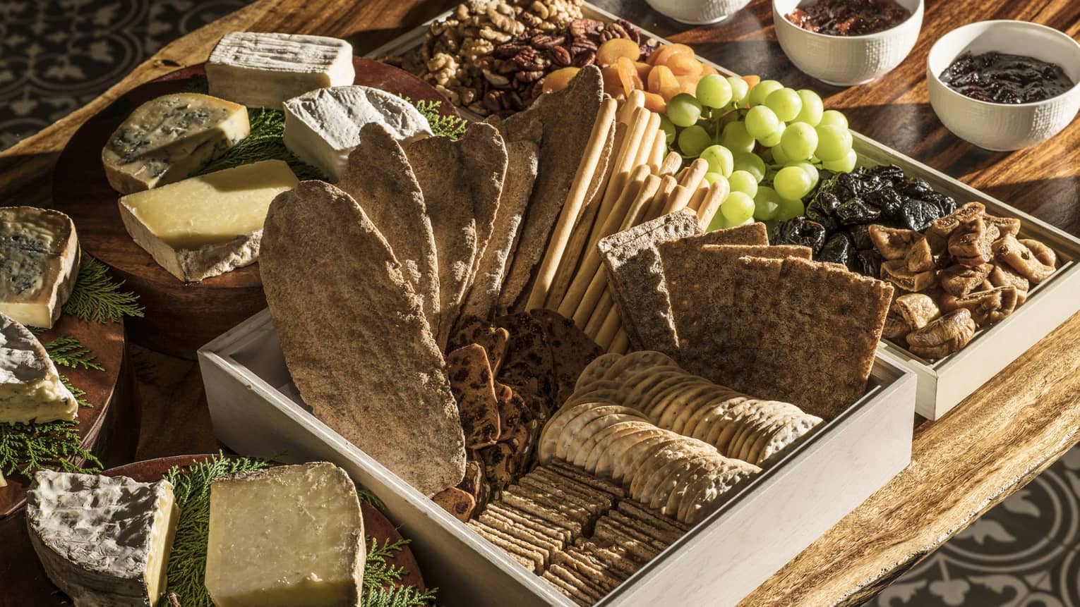 A charcuterie board with crackers and bread, a variety of cheeses, fresh and dried fruit, jams and nuts