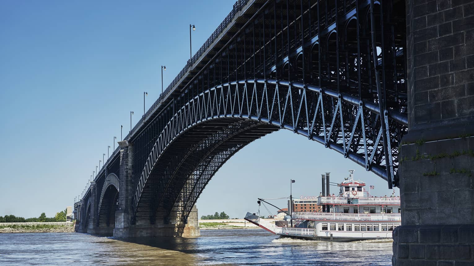 A river boat going under a large metal bridge on a bright day on a river.