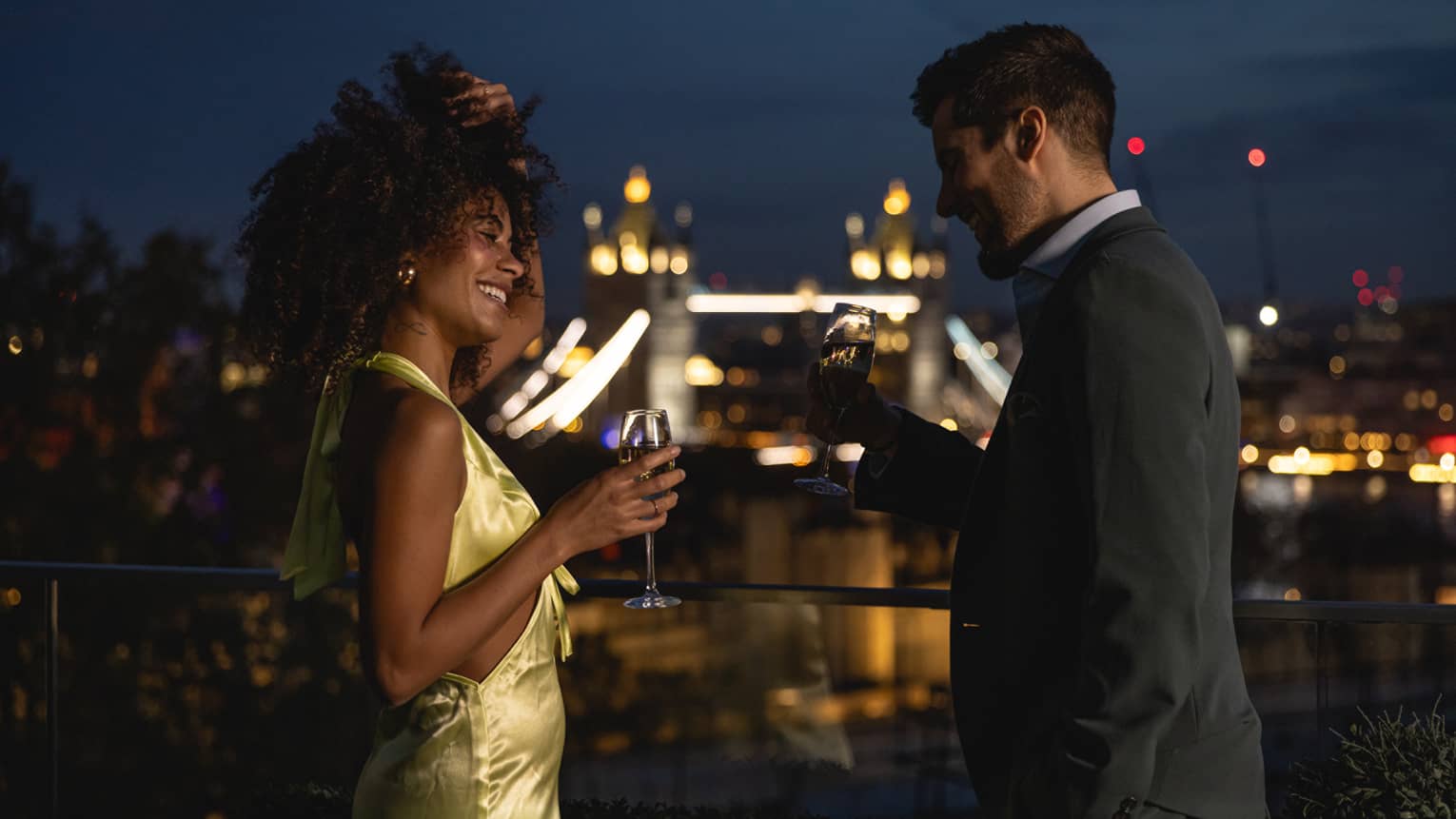 Couple standing on terrace, laughing with champagne in hand over London views at night
