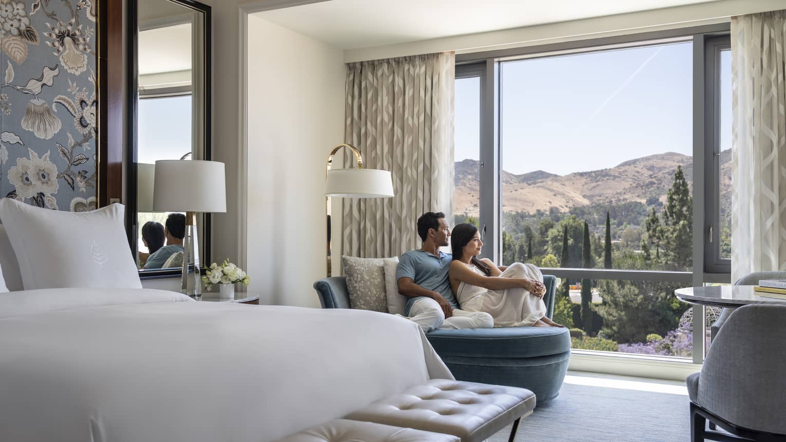A couple lounging on a chair in their hotel room looking out at the mountains.