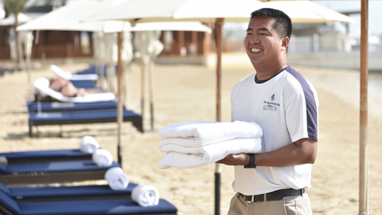 Beach attendant wearing white t-shirt and khaki pants smiles as he carries two fresh towels past navy-blue lounge chairs on the beach