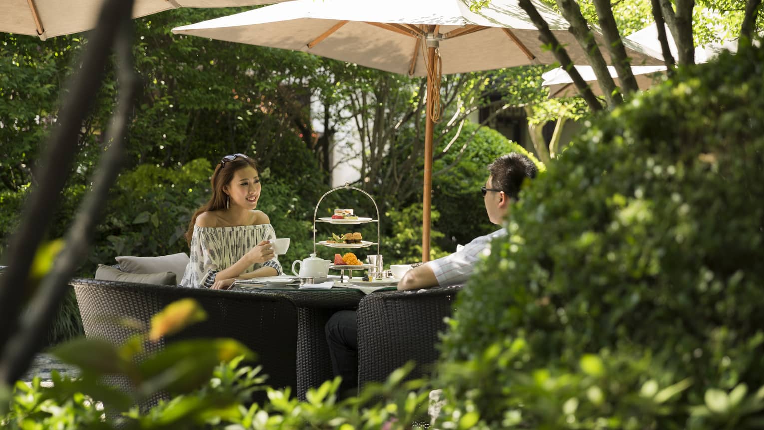 Woman and man drink tea near tray of sweets at table under shade of patio umbrella