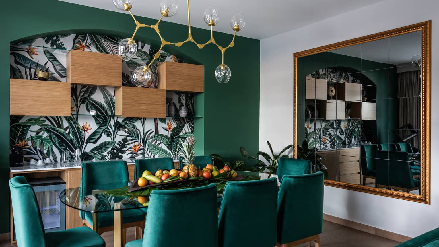 A dining area with emerald green chairs, a jungle mural and an Art Deco chandelier.