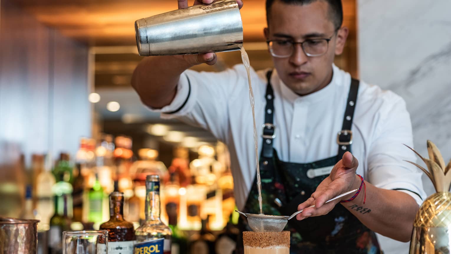 A man in a Four Seasons apron pours a drink through a sifter into a tall glass.