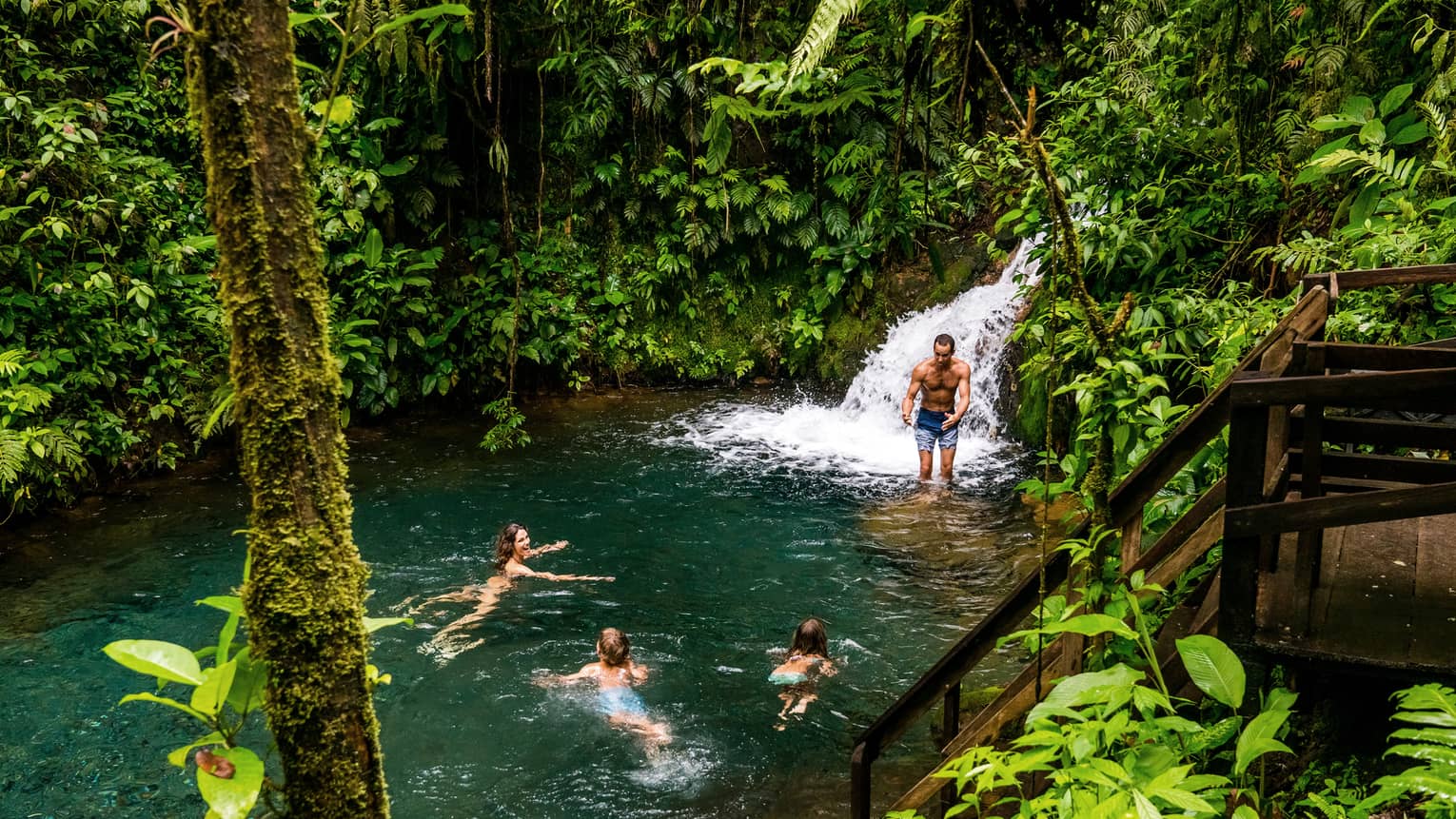Two adults and two kids frolic at the foot of a waterfall surrounded by lush jungle greenery