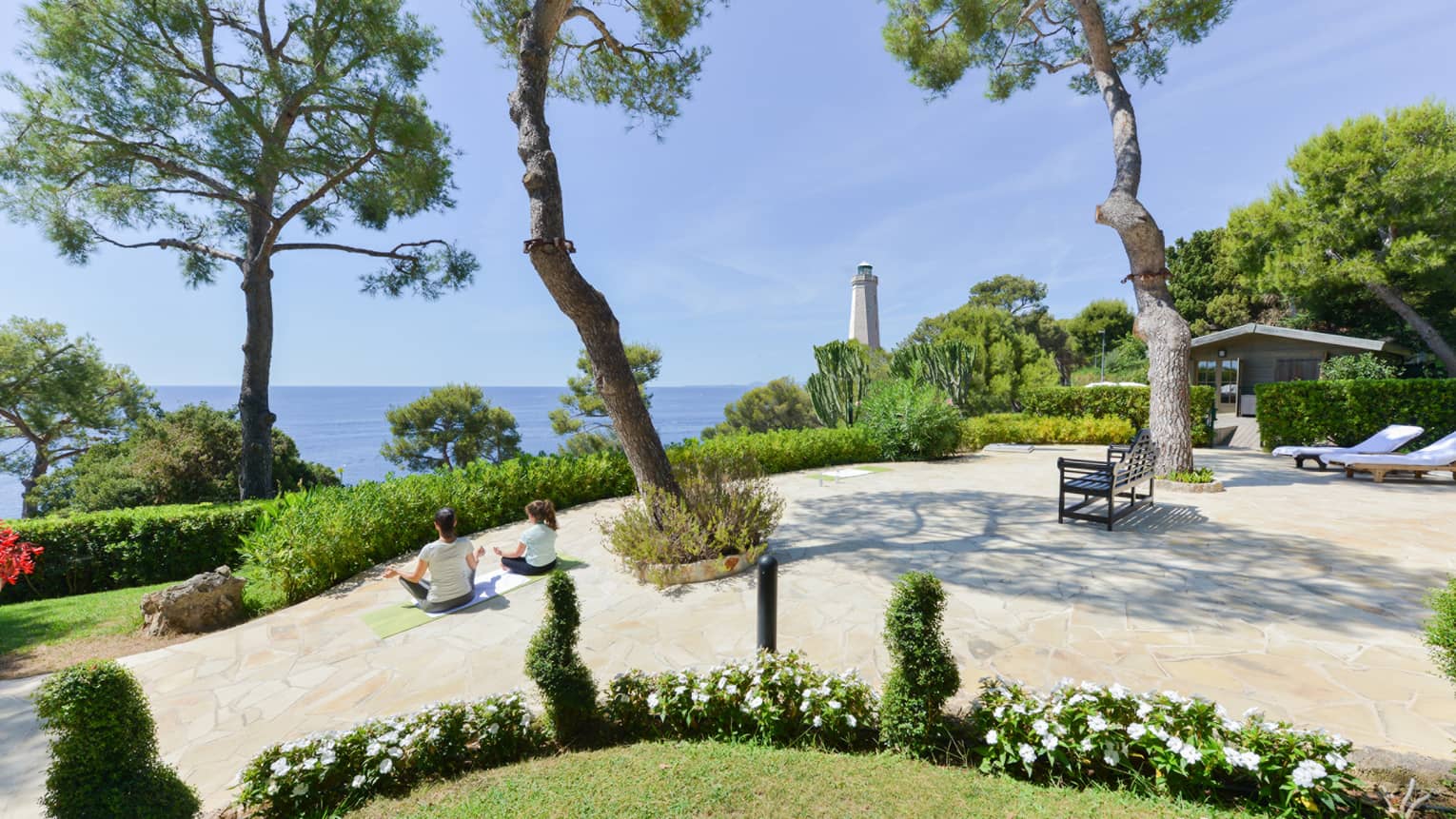 Rear view of two people in a seated yoga pose on curved patio with lush greenery, sea and lighthouse view