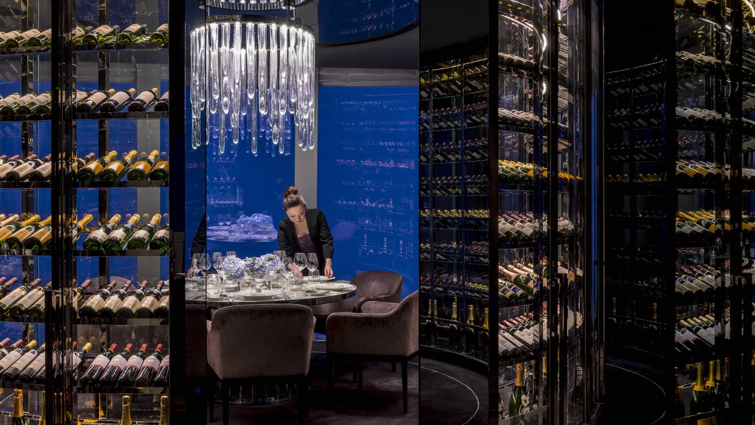 Woman sets plate on round private dining table under crystal chandelier, surrounded by wine cellars