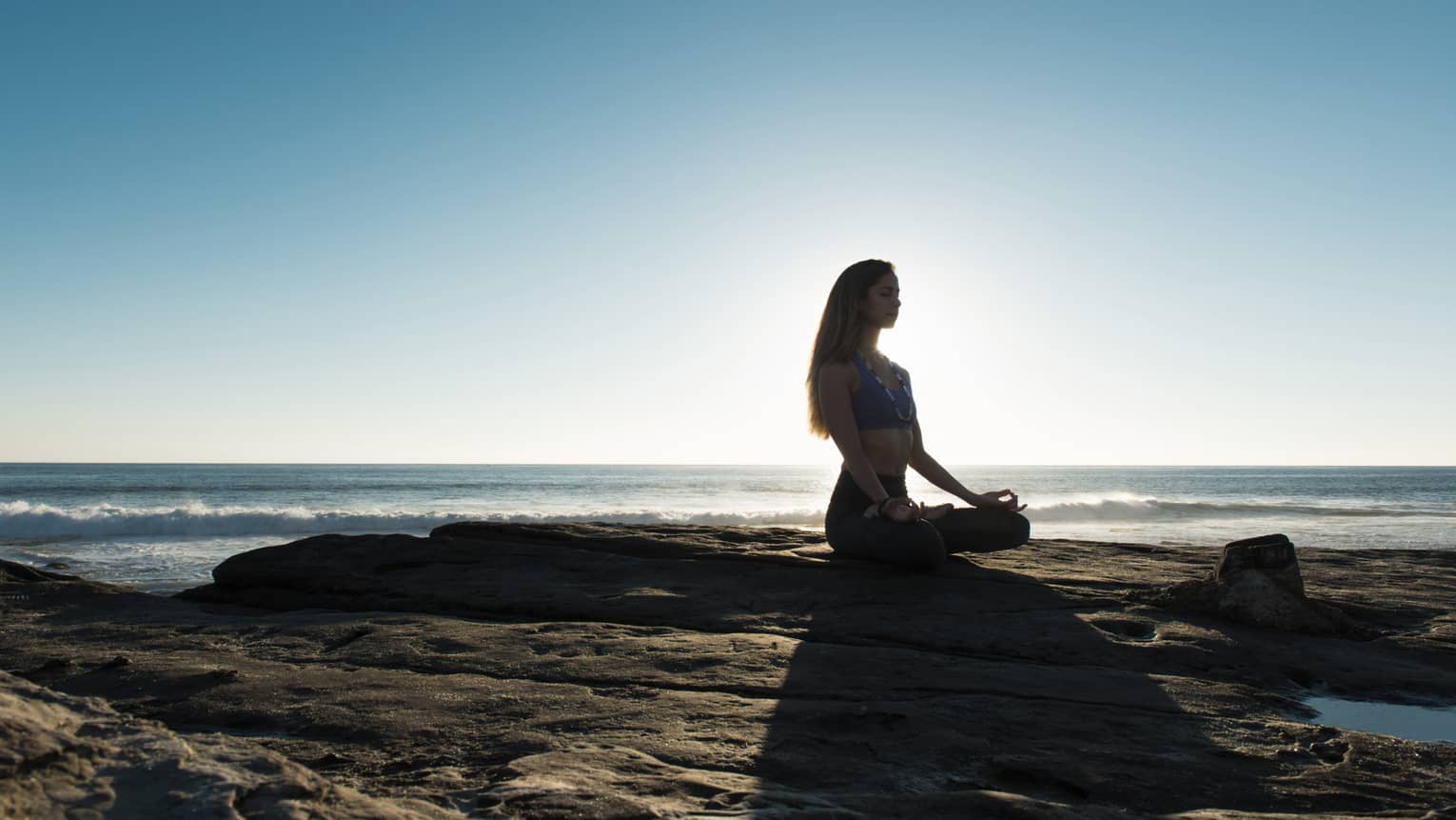 Silhouette of woman sitting cross-legged, meditating on rock by ocean