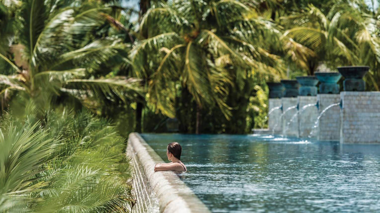 Woman wades, looks out from edge of outdoor swimming pool with fountains