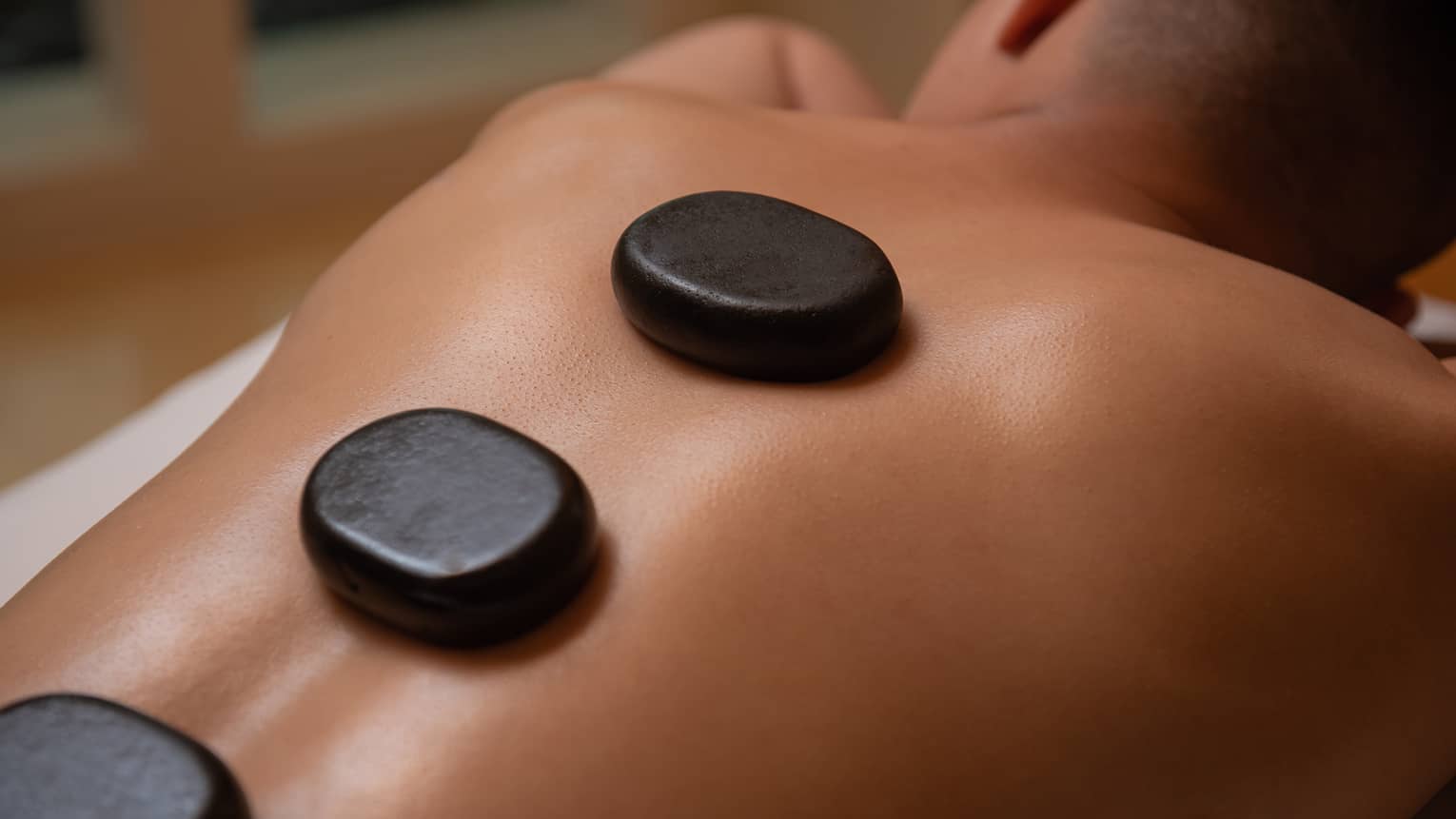 Three large black stones rest on man's bare back and spine as he lays on massage table