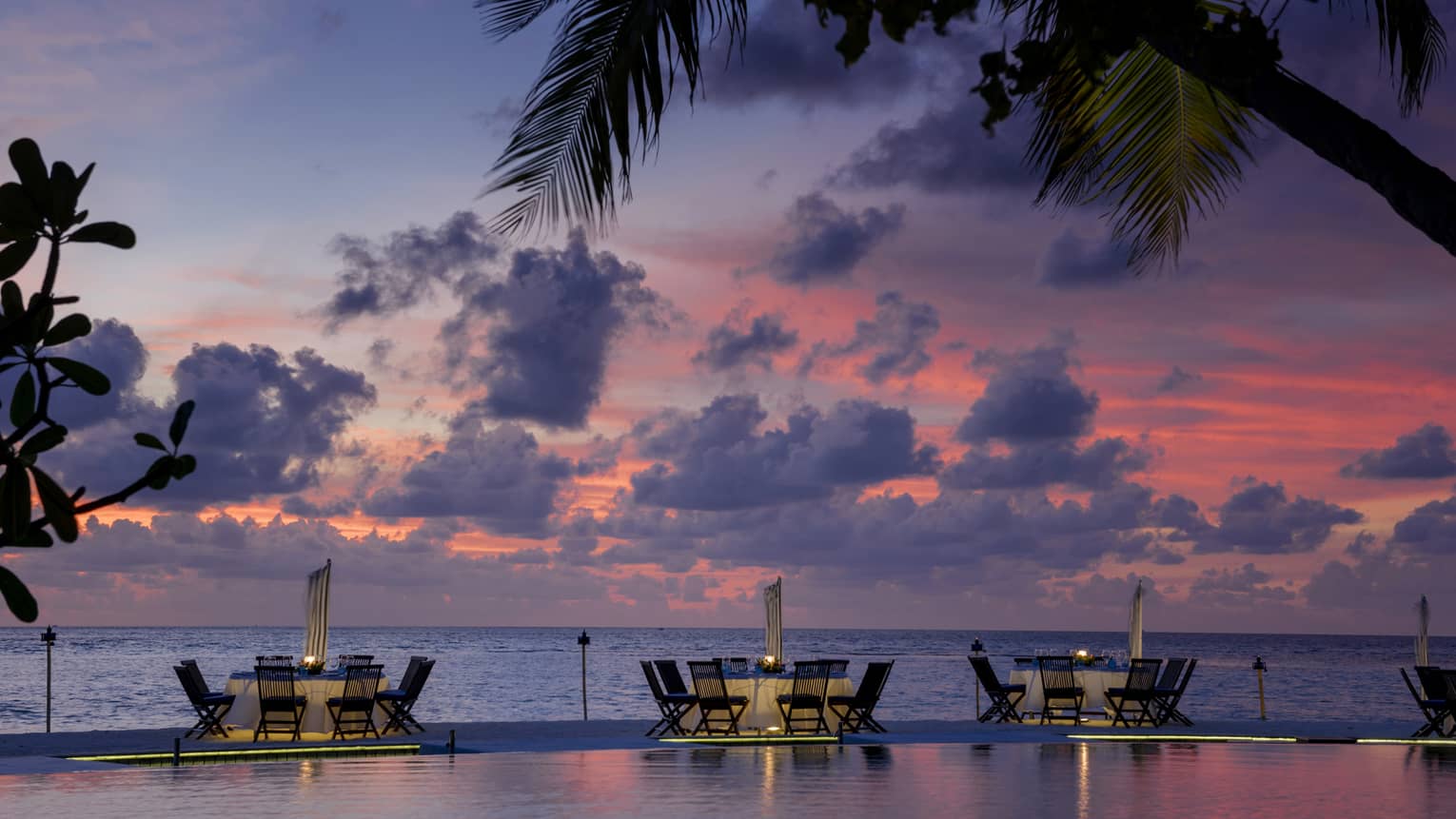 Tables with chairs between infinity pool and ocean at dusk with palm tree