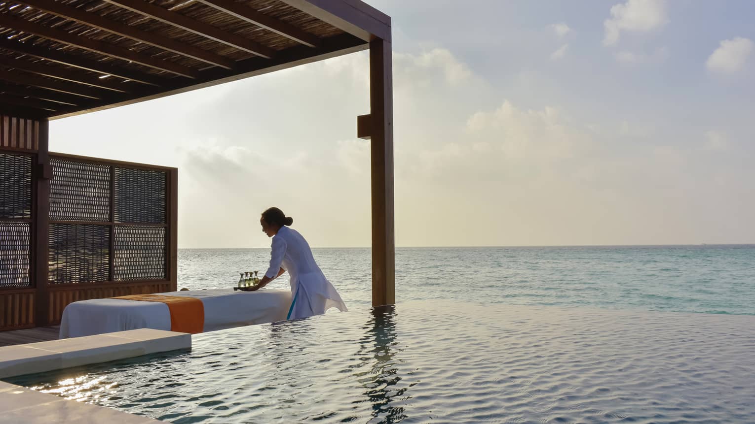 Woman sets tray down on table in front of infinity pool alongside ocean