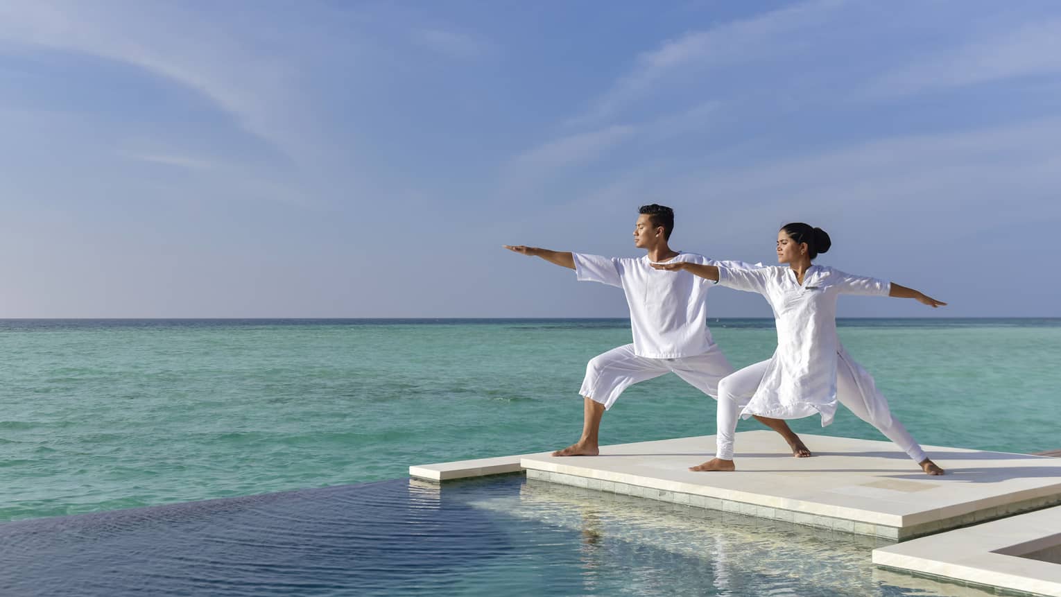 A couple dressed in all white does yoga on a white dock over the sea