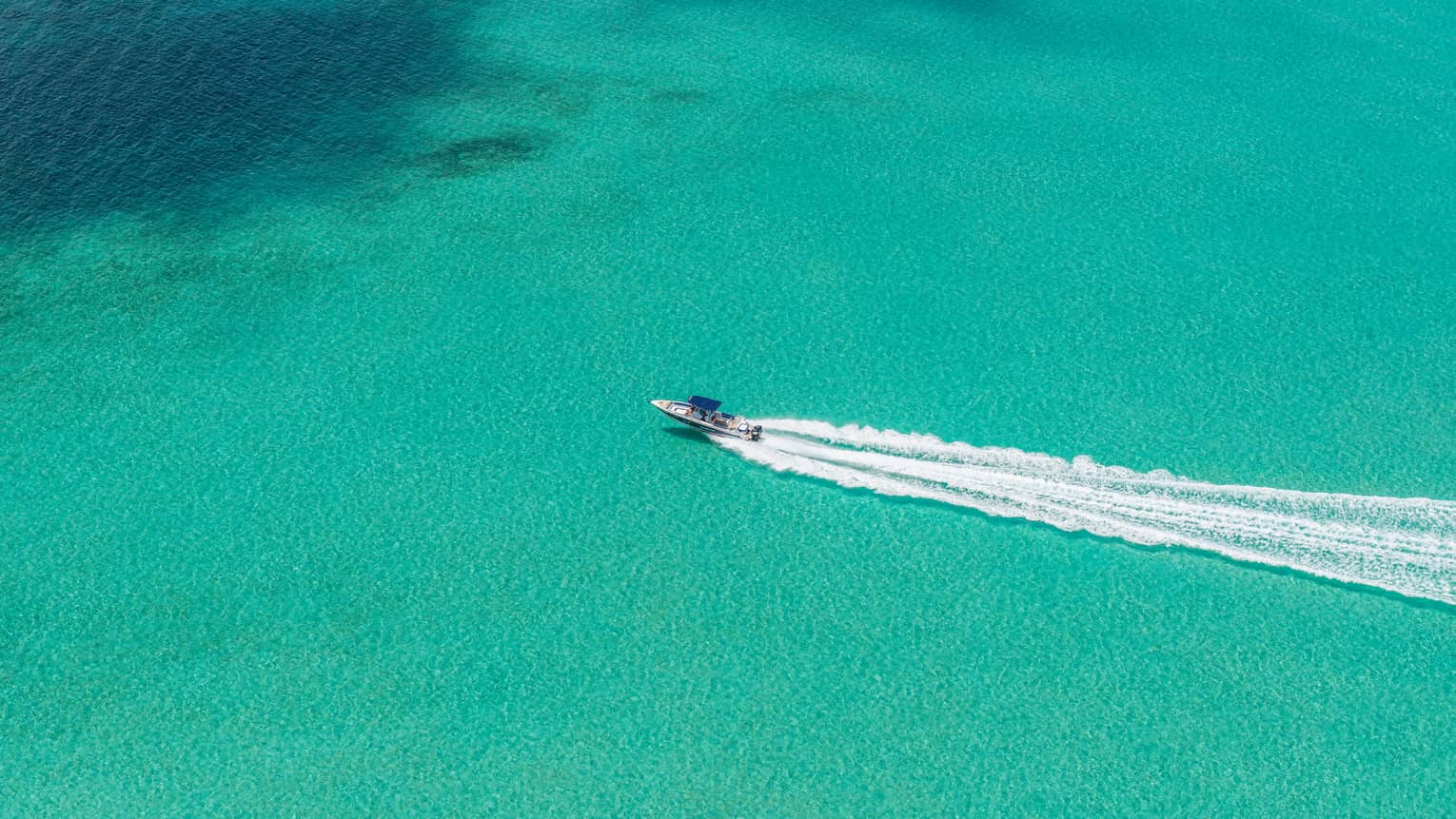 Aerial view of a speedboat moving through a vast expanse of turquoise water, followed by a substantial wake trail.