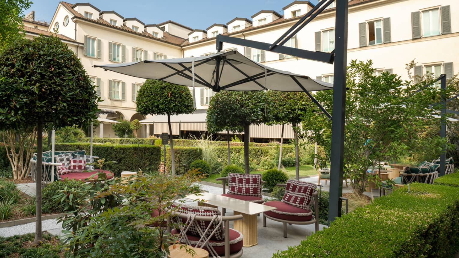 Elegant courtyard vignette under tan umbrella surrounded by hedge and topiaries with hotel in backdrop