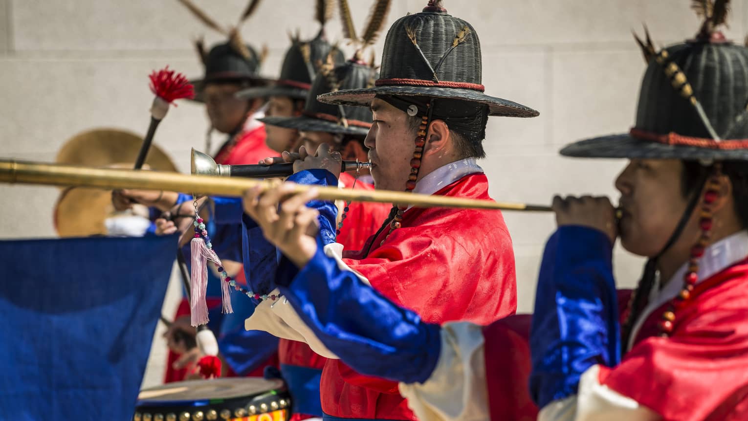 Profile of men in decorated hats and blue-sleeved red satin smocks playing ancient Korean wind and percussion instruments.