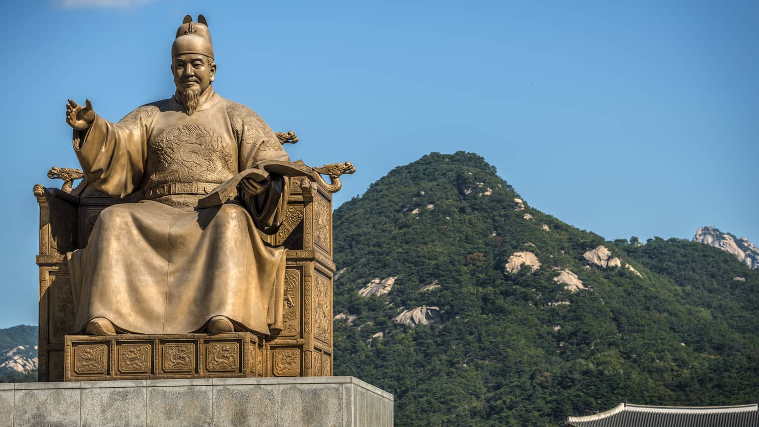 Golden statue of Sejong the Great, cloudless blue sky, lush green mountains in the background