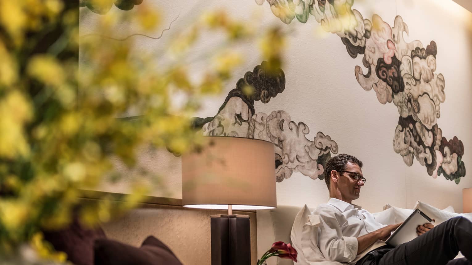 Man lounges, reads iPad on sofa next to lamp under black-and-white Chinese brushwork mural