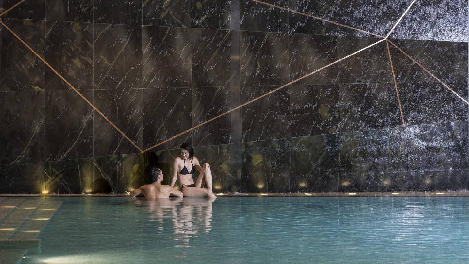 Woman sits on edge of indoor swimming pool, man wades beside her 
