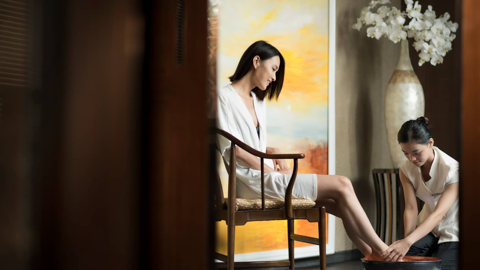 Spa staff rinses woman's feet in bowl as she sits in chair in white robe