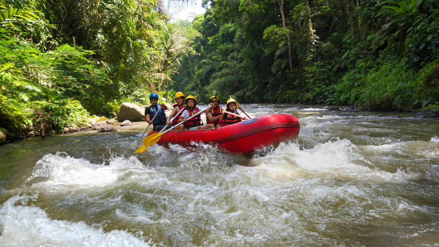Family of four and guide hold paddles, steer red inflatable raft on rapids in river