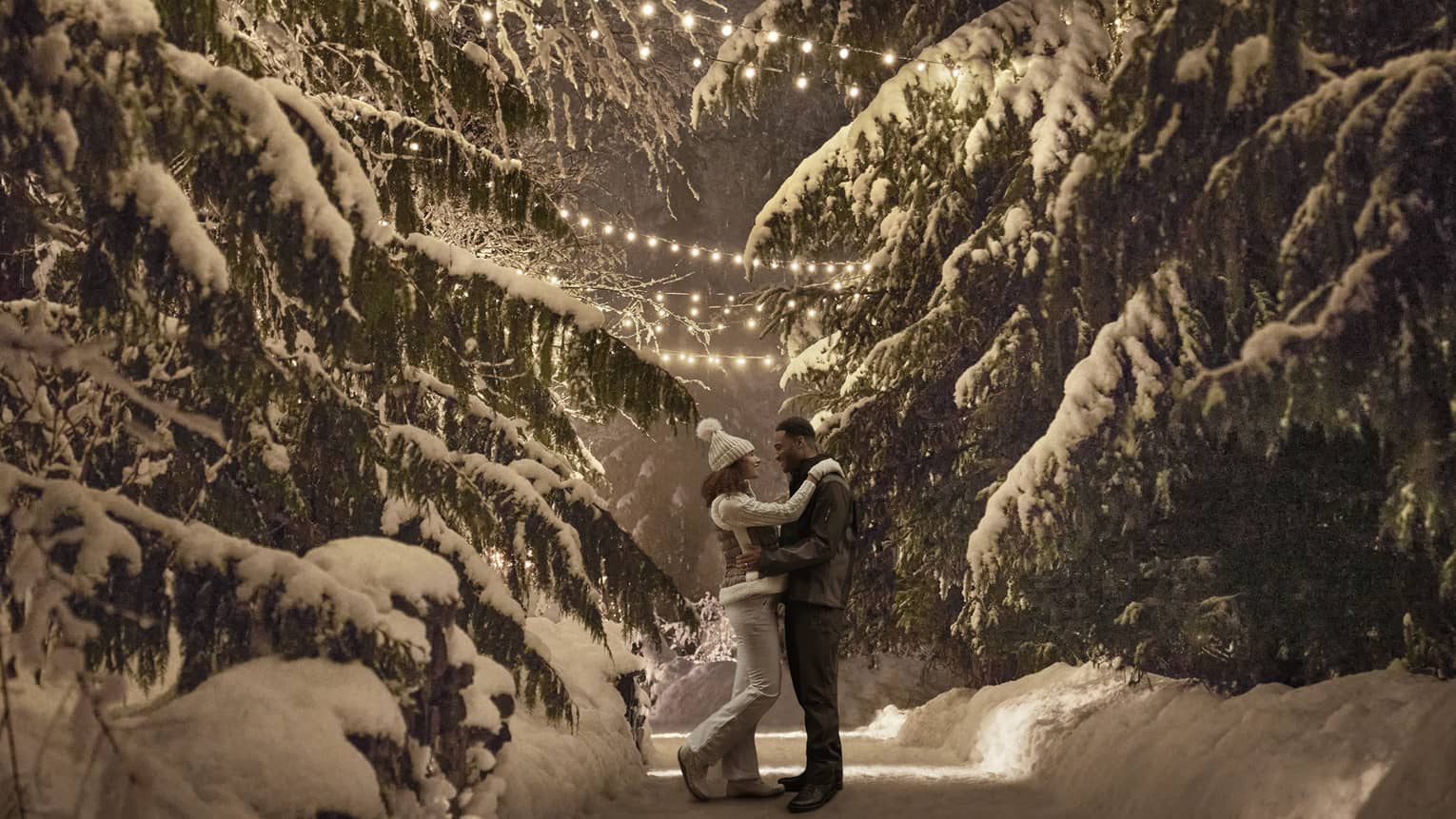 Couple wearing winter coats embrace on snow covered path between large trees at night