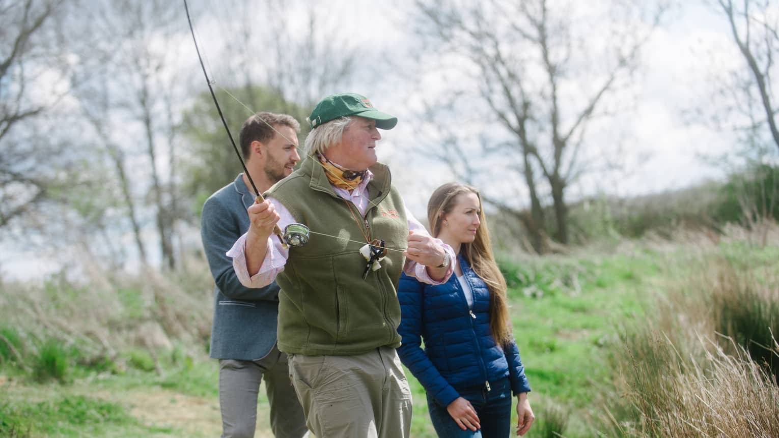 An older man and a man and woman fishing outside surrounded by thin trees and tall grass.