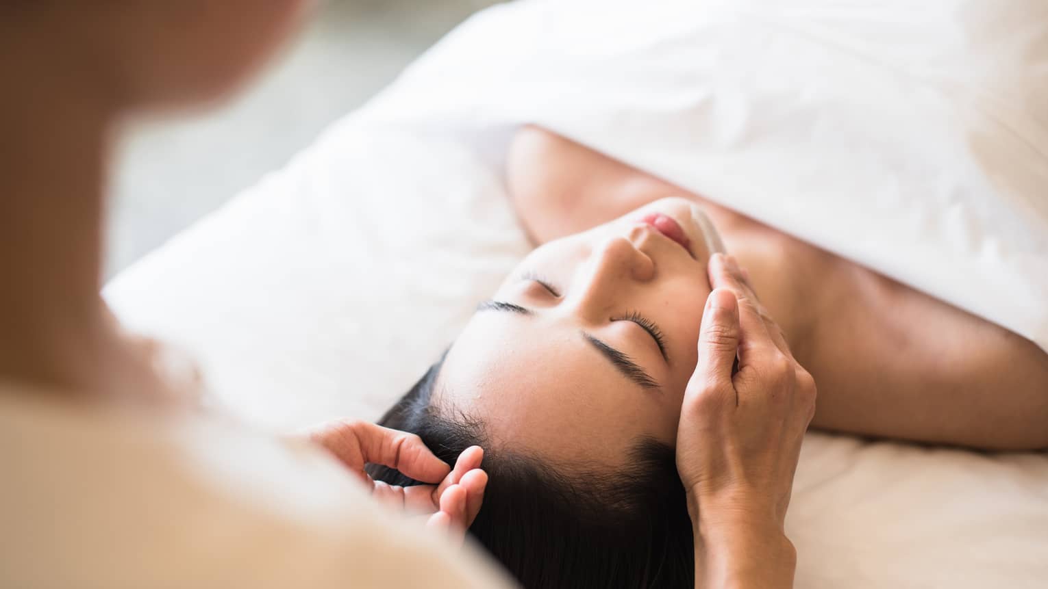 Woman lying under white sheet on massage table, eyes closed as spa staff rubs lotion on her chin