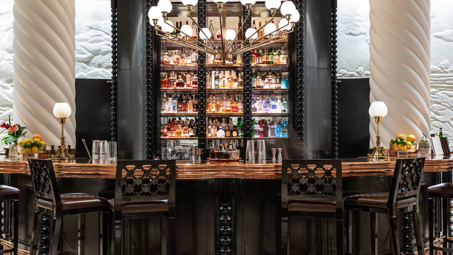 Wall of liquor bottles behind the wooden bar, four black barstools in front, white columns on either side
