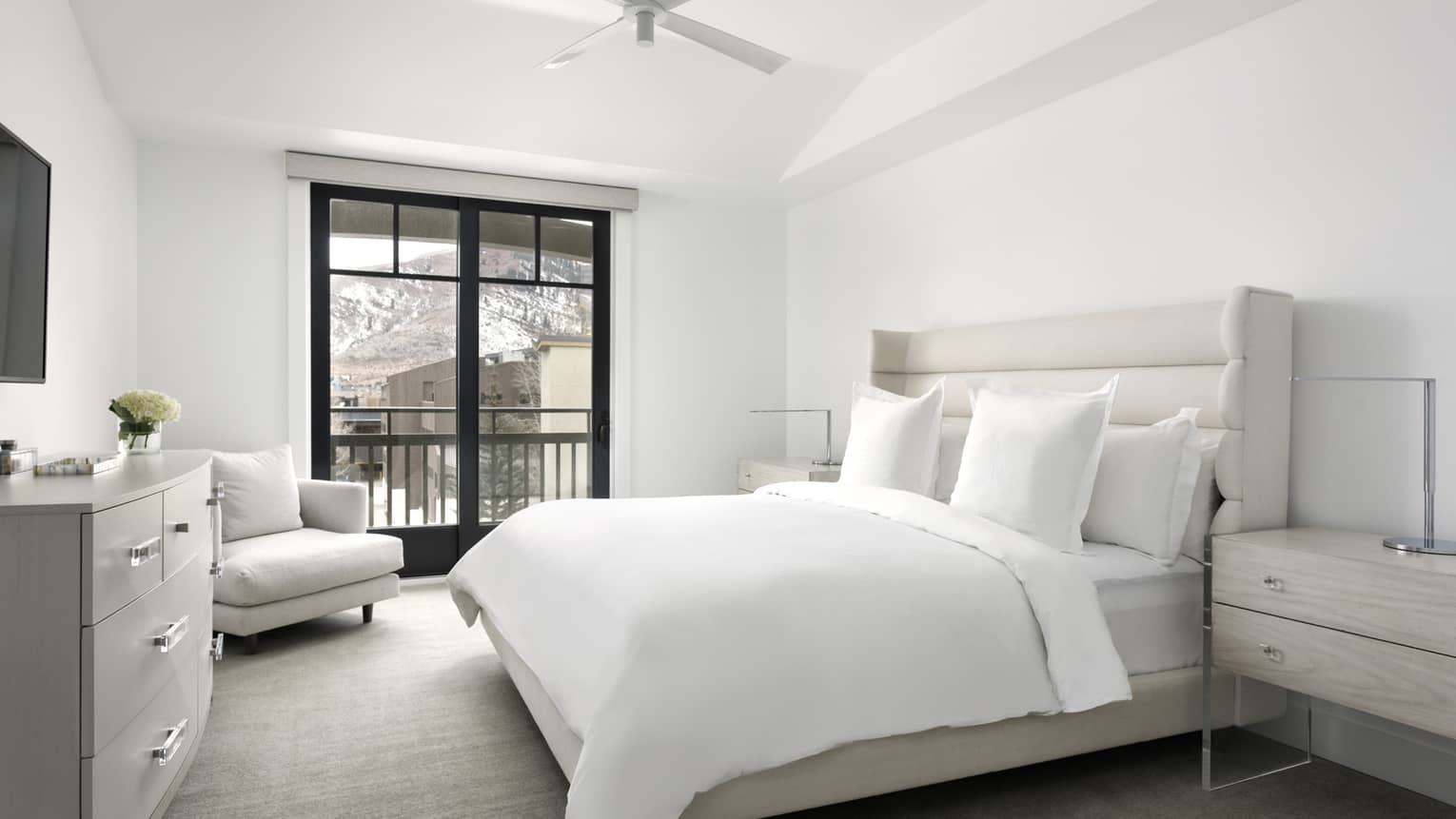 Bedroom with white walls, white king bed, light grey arm chair, dresser, walk-out balcony