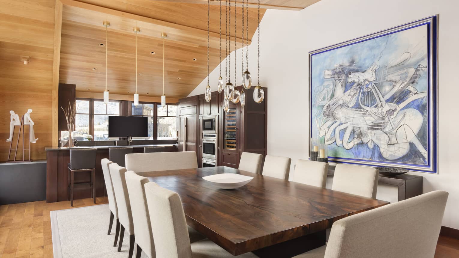 Dining table with 10 cushioned chairs, leading into kitchen with wooden vaulted ceiling
