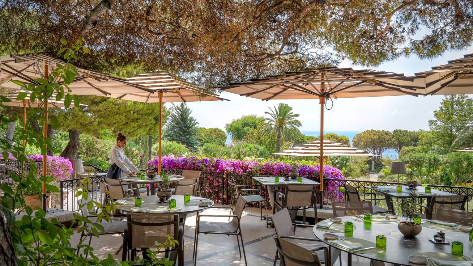 Waiter sets a table on outdoor restaurant terrace, surrounded by greenery and magenta-coloured flowers