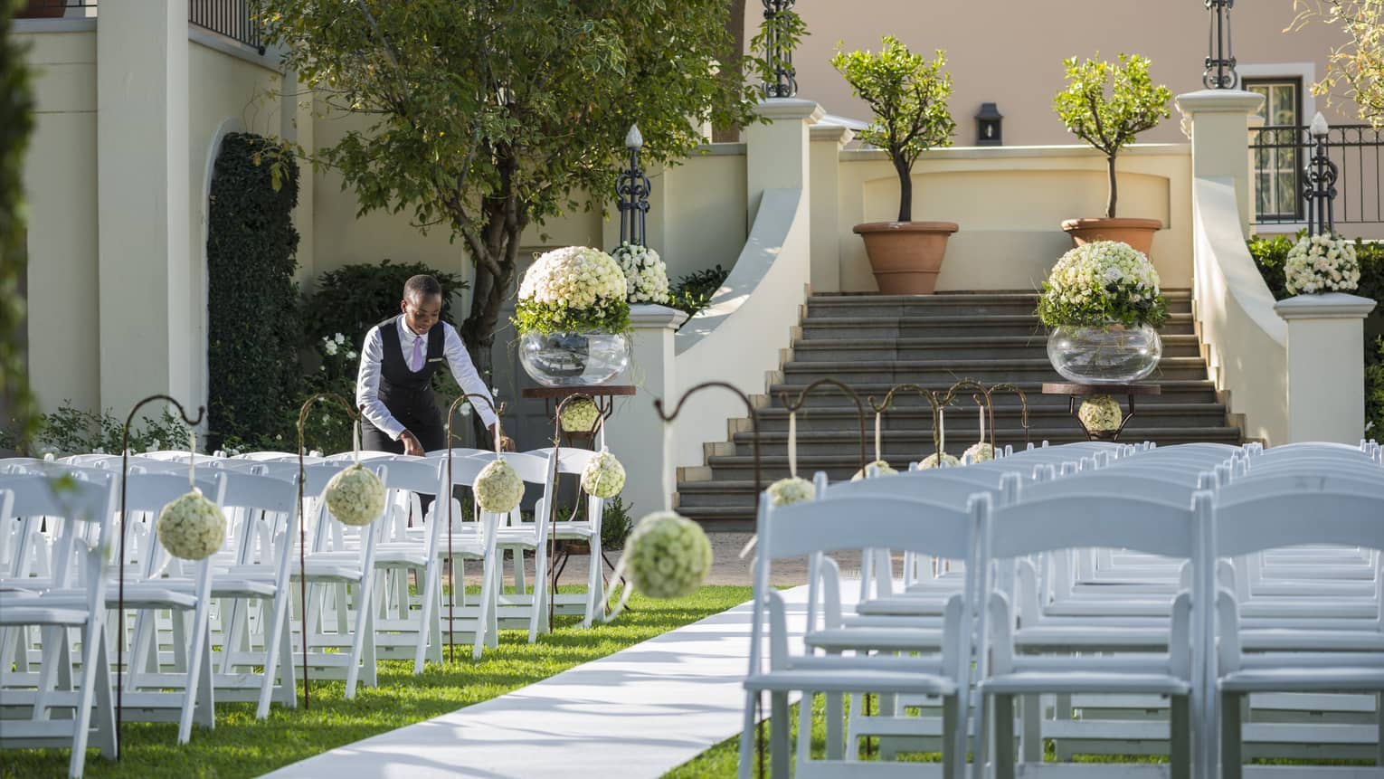 Hotel staff arranges white chairs on garden lawn for wedding service 
