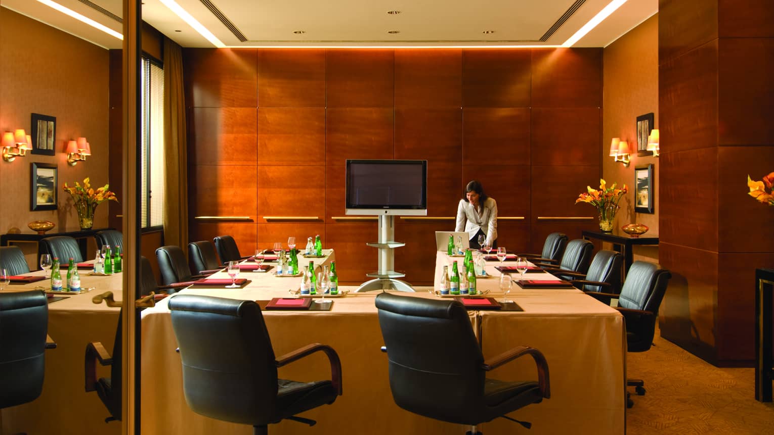 Woman stands in front of U-shaped meeting table set with folders, bottles of water, lined with black office chairs