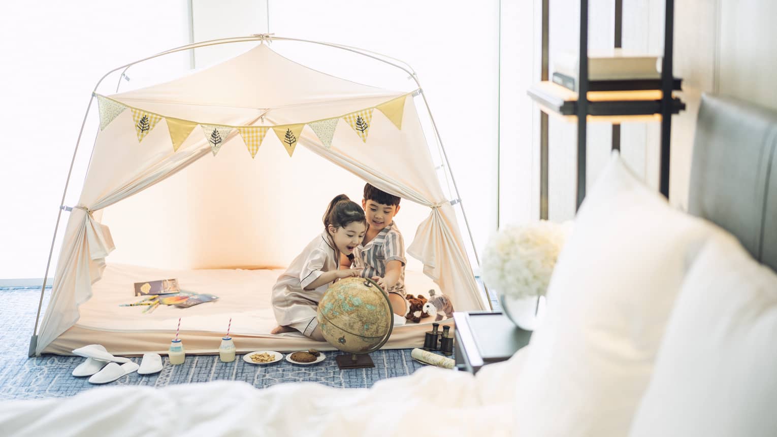 Two smiling children sit in a white play tent in a guest room at Four Seasons Hotel Seoul, with toys and snacks around them