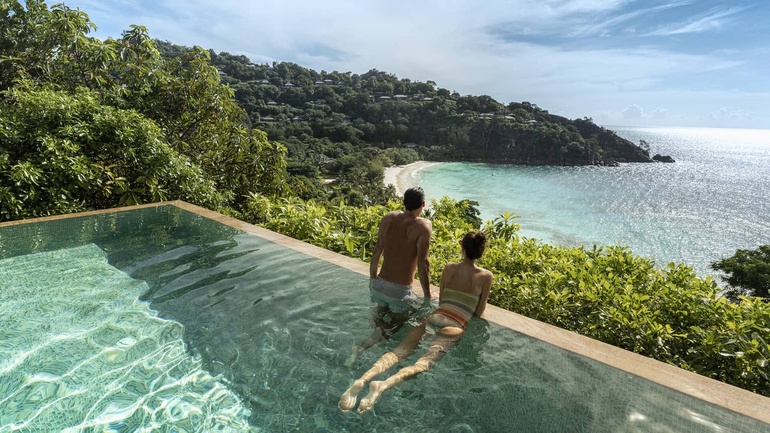 A couple looks out at a view of crystal blue waters and cliffs from an infinity pool