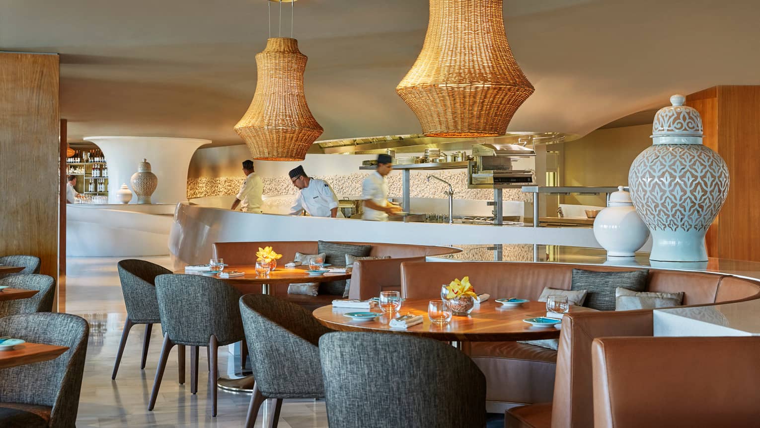 Large wicker lamps hang over mid-century style dining tables and chairs at Blue restaurant, chefs work in background