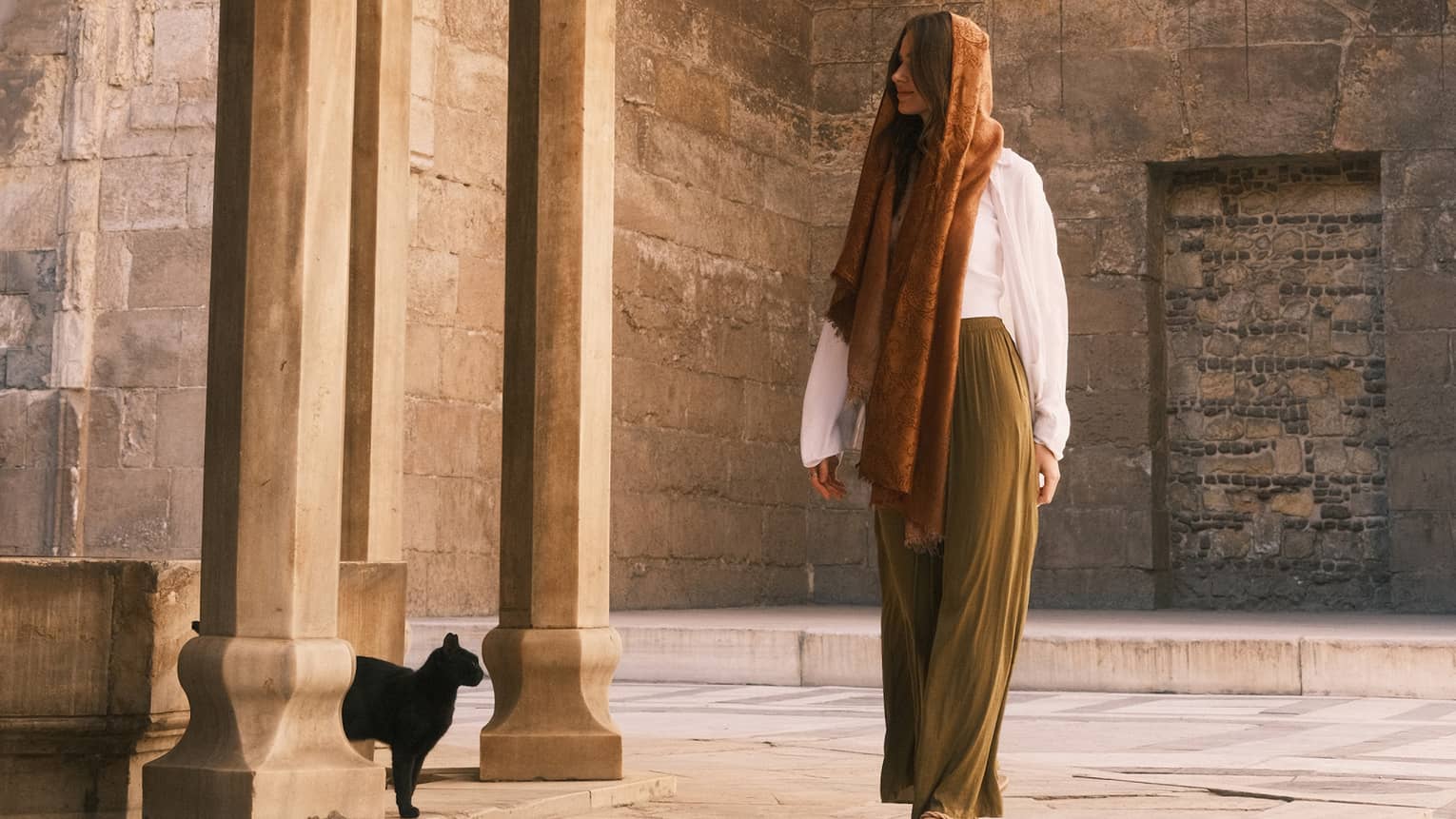 A person, head covered, stands under a pyramidal mosque lantern amid ornately carved walls and columns, a black cat nearby.