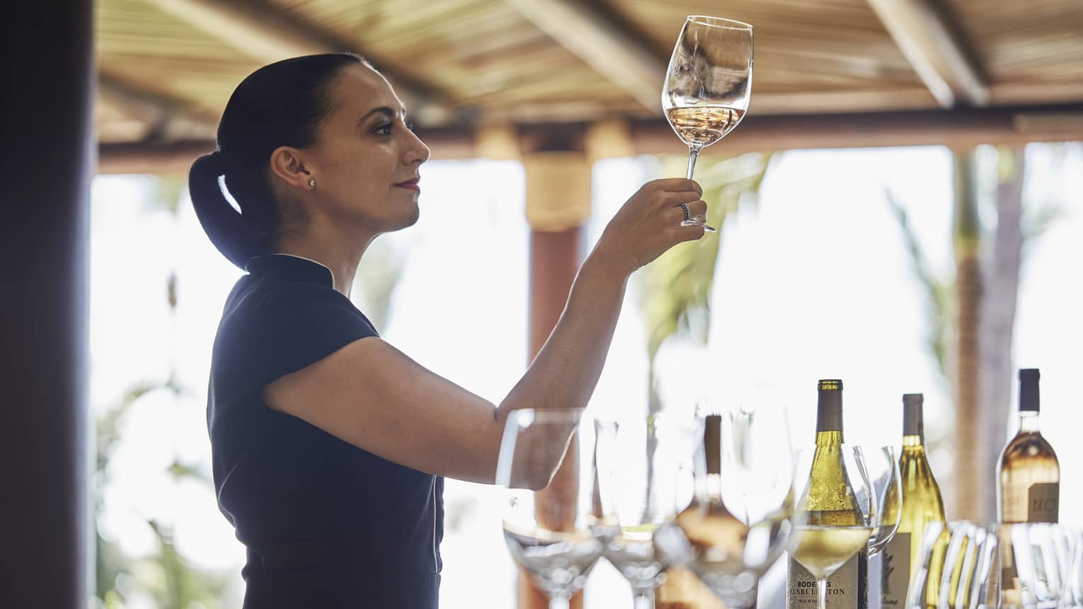 Sommelier studies a glass of pale wine held at eye level, other bottles and empty glasses spread out on the bar counter. 