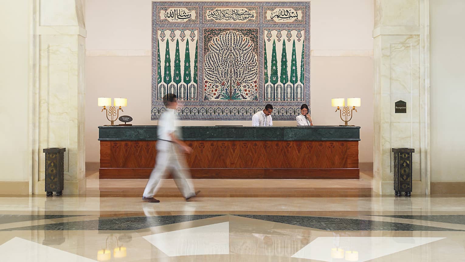 Man walks through hotel lobby, past staff at reception desk under large tile mosaic