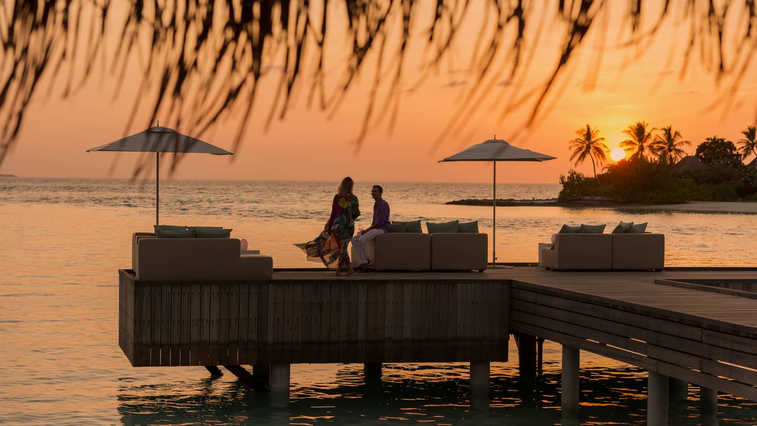 Private island dining at sunset. Woman with long dress blowing in the wind, man sits on the edge of white sofa