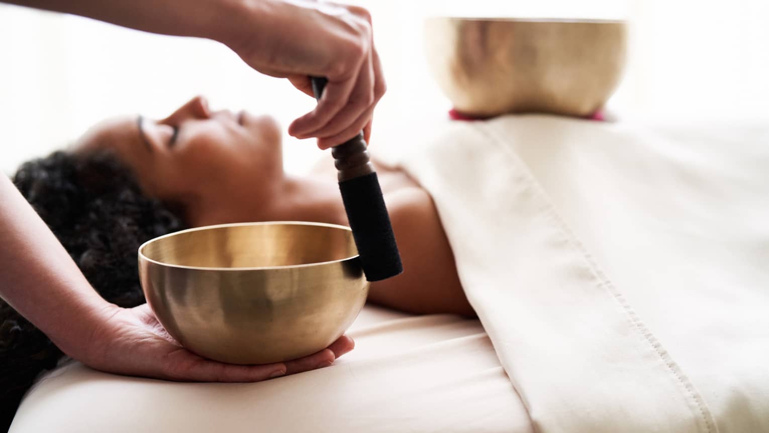 Woman with eyes closed lies on massage table in as vibrational sound therapy is performed with a Tibetan singing bowl